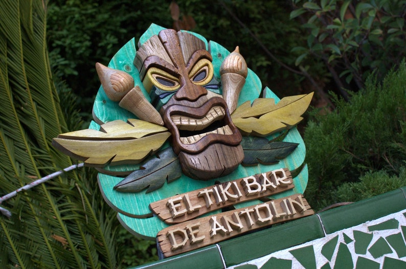 May include: A wooden tiki mask with carved details and a green background. The sign reads "El Tiki Bar de Antolin".