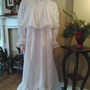 May include: A long, white nightdress with a high collar, long sleeves, and a ruffled hem. The nightdress features lace trim on the sleeves and a decorative yoke. A pink flower is at the neckline. The nightdress is displayed on a mannequin.
