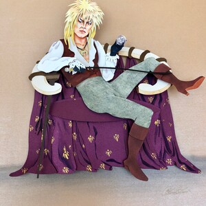 May include: A papercraft figure of a character with long blonde hair, wearing a brown and white costume, sitting on a purple and gold throne. The character is holding a small orb in one hand and a staff in the other.