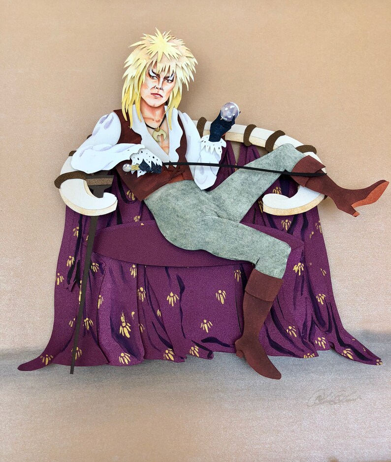 May include: A papercraft figure of a character with long blonde hair, wearing a brown and white costume, sitting on a purple and gold throne. The character is holding a small orb in one hand and a staff in the other.