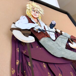 May include: A papercraft sculpture of a character from the movie Labyrinth. The character is sitting on a throne with a purple and gold patterned fabric. The character is wearing a white shirt, a brown vest, and grey pants. The character has blonde hair and is holding a sword.