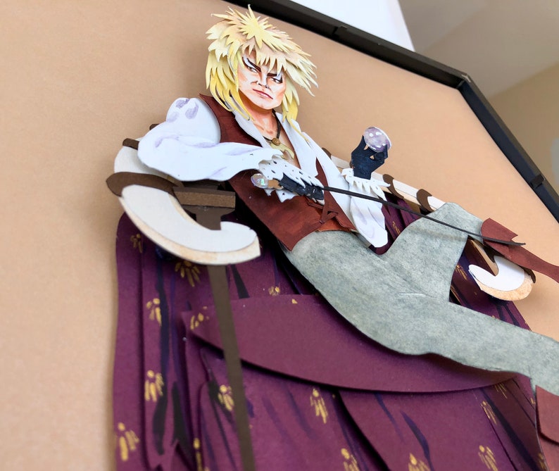 May include: A papercraft sculpture of a character from the movie Labyrinth. The character is sitting on a throne with a purple and gold patterned fabric. The character is wearing a white shirt, a brown vest, and grey pants. The character has blonde hair and is holding a sword.