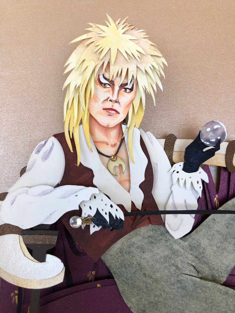 May include: A papercraft figure of a character with long, blonde hair, wearing a white shirt, brown vest, and grey pants. The character is holding a crystal ball in one hand and a sword in the other.