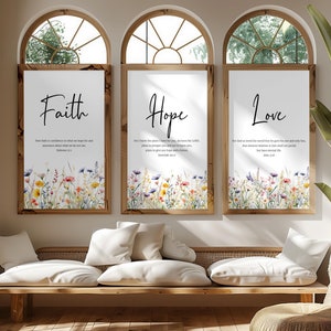 May include: Three framed art prints with the words "Faith", "Hope", and "Love". Each print has a watercolour floral border. The artwork is displayed in a bright room with natural wood accents. The prints are in a room with arched windows.