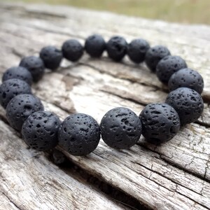 May include: A black lava rock beaded bracelet with a rough, textured surface. The bracelet is made of round beads and is lying on a piece of weathered wood.
