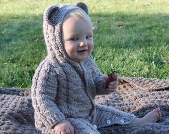 May include: A baby wearing a brown crocheted hooded jumpsuit with bear ears. The baby is sitting on a brown blanket and holding a small brown leaf.
