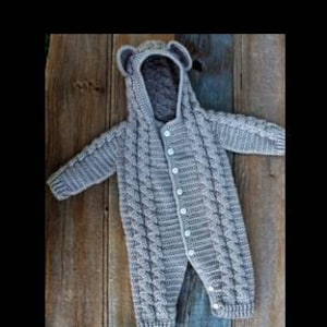 May include: A gray knitted baby jumpsuit with a hood featuring bear ears. The jumpsuit has a cable knit design and is buttoned down the front. It is displayed on a weathered wooden surface.