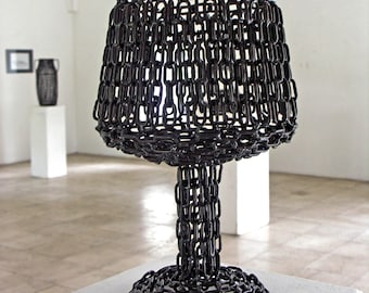 May include: A black metal sculpture of a wine glass, made entirely of linked chains. The sculpture is displayed on a white pedestal.