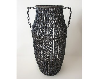 May include: A tall, black metal vase made entirely of linked chains. The vase has two handles and a rounded base.