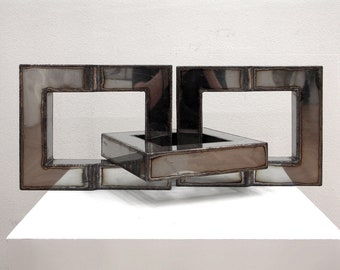May include: A metal sculpture made of three interlocking squares. The squares are made of polished metal and have a reflective surface. The sculpture is on a white pedestal.