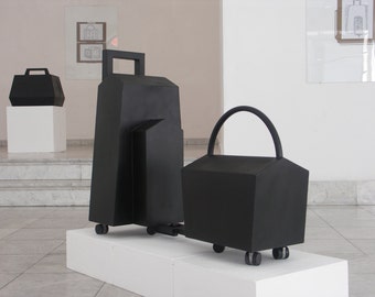 May include: Three black, geometrically shaped, wheeled containers. The largest container has a handle and is in the center. The smallest container has a handle and is on the right. The middle container is attached to the largest container.