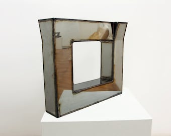 May include: A silver-colored metal sculpture in the shape of a square frame with a smaller square frame inside. The sculpture is on a white pedestal.