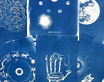 May include: A collection of five cyanotype prints on paper featuring white ink illustrations. The prints display celestial themes including a star chart, lunar phases, a full moon, a zodiac wheel, a palmistry hand, and a cat with a sunburst.