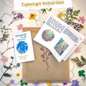 May include: A brown paper envelope with a small blue booklet titled "CyanoLab Instructions" and a folded white booklet with instructions for creating cyanotype prints. The envelope is surrounded by dried flowers and leaves on a white lace tablecloth.