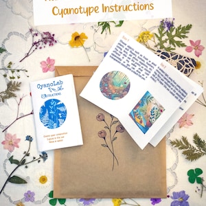 May include: A brown paper envelope with a small blue booklet titled "CyanoLab Instructions" and a folded white booklet with instructions for creating cyanotype prints. The envelope is surrounded by dried flowers and leaves on a white lace tablecloth.