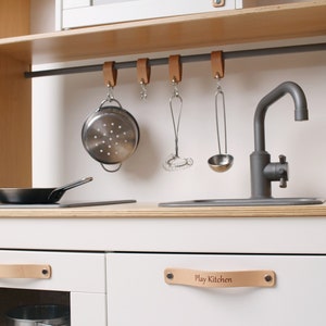 May include: A wooden play kitchen with white cabinets and a light wood countertop. It features a stainless steel sink and faucet, a hanging rail with cooking utensils, and a small frying pan. The drawer handles are leather with the text "Play Kitchen".
