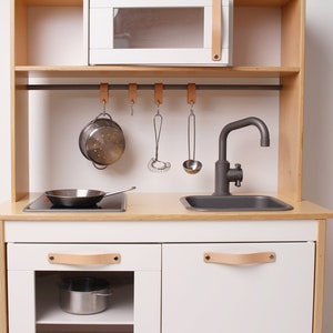 Pode incluir: Uma cozinha de brinquedo de madeira branca com uma pia e uma torneira pretas, um fogão preto e um forno de microondas. A cozinha tem dois armários com puxadores de couro e uma prateleira acima do fogão. Há ganchos acima do fogão com um escorredor, um batedor e uma concha pendurados.