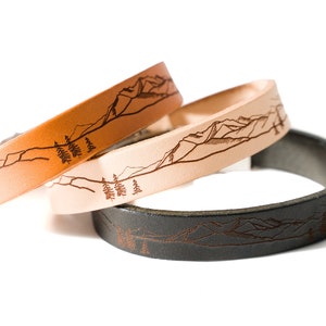 Puede incluir: Tres pulseras de cuero con un diseño de cordillera montañosa y pinos. Las pulseras son de color marrón, beige y negro.