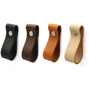 Könnte beinhalten: Vier Leder-Schrankgriffe in Schwarz, Braun, Beige und Creme mit einem silbernen Metallknopf.