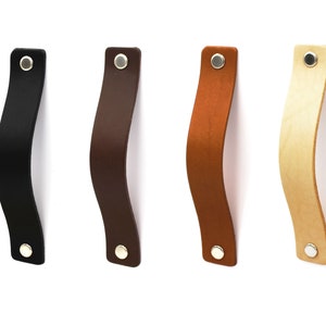 Könnte beinhalten: Vier Leder-Schubladengriffe in verschiedenen Farben: Schwarz, Braun, Beige und Natur. Jeder Griff hat eine silberne Schraube.