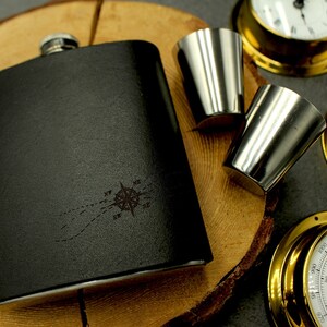 Könnte beinhalten: Schwarze Flasche mit Kompassrosen-Design und zwei Schnapsgläsern aus Edelstahl. Die Flasche ist rechteckig und hat eine strukturierte Oberfläche. Dekorative nautische Instrumente im Hintergrund.
