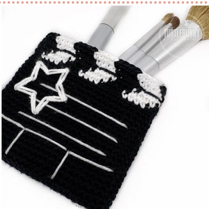 May include: A black and white crochet makeup bag with a star and film strip design. The bag is filled with makeup brushes. The text "Hollywood Makeup Bag" is written below the bag.