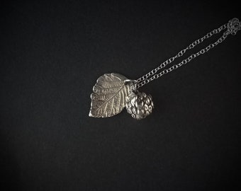 May include: A silver necklace with a pendant featuring a detailed leaf and a berry. The pendant is attached to a silver chain.
