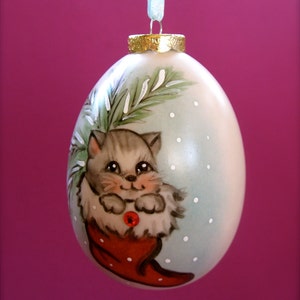 May include: A white Christmas ornament with a hand-painted design of a gray cat wearing a red stocking. The cat is surrounded by green pine branches and white snowflakes.