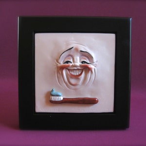 Puede incluir: Un azulejo de cerámica blanco con una cara esculpida en 3D sonriendo y sosteniendo un cepillo de dientes con pasta de dientes azul. El azulejo está enmarcado en negro.