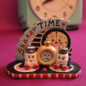 May include: A ceramic kitchen figurine with the words "Shaker Time" features two salt shakers and a clock. The salt shakers have smiling faces and red shoes. A vintage mint green clock is in the background.