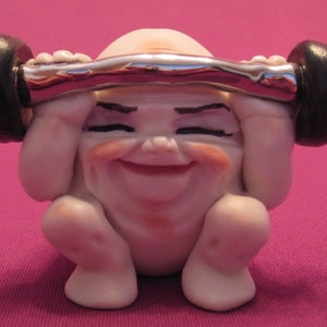 May include: A white ceramic figurine of a smiling person lifting a silver barbell with black weights. The figurine is a whimsical representation of a person exercising.