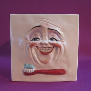 May include: A white ceramic wall plaque with a 3D sculpted face smiling with a large toothy grin. The face has exaggerated features and a red toothbrush with blue toothpaste is placed in front of the mouth.