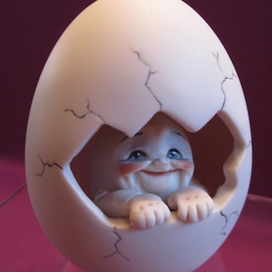 May include: A white ceramic egg figurine with a cracked surface, revealing a smiling baby doll inside. The baby doll has blue eyes and rosy cheeks.