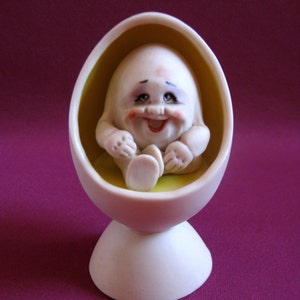Puede incluir: Una figurita de cer&aacute;mica blanca de un beb&eacute; sonriente sentado en una silla en forma de huevo amarilla. La silla tiene una base blanca.