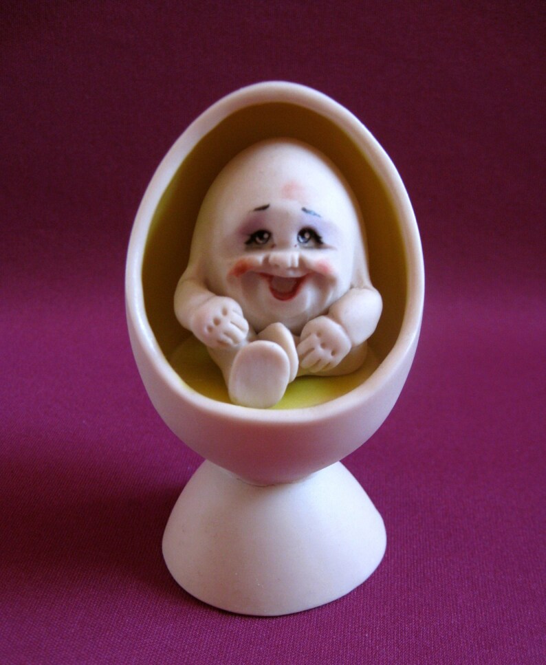 Puede incluir: Una figurita de cer&aacute;mica blanca de un beb&eacute; sonriente sentado en una silla en forma de huevo amarilla. La silla tiene una base blanca.