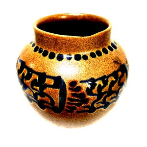 May include: A small, round ceramic vase with a wide opening. The vase is primarily tan with dark blue decorative patterns and a band of blue dots near the rim. The interior is dark brown.