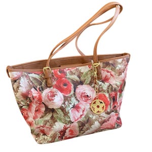 May include: Floral print tote bag with brown leather handles and trim. The bag features a pattern of red and pink roses with green leaves. A gold-colored charm hangs from the handle. The bag has a rectangular shape.