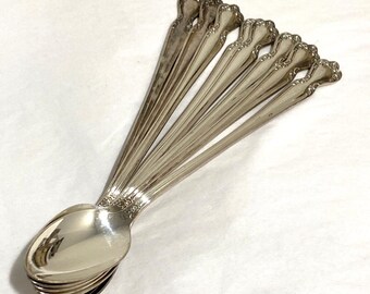 May include: A collection of silver spoons arranged in a fan shape. The spoons have a decorative floral design on the handles and a shiny, reflective surface. The spoons are likely vintage or antique, and are set against a white background.