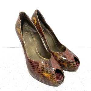 Op de afbeelding: Een paar Stuart Weitzman peep-toe pumps. De schoenen hebben een slangenprint in bruin-, rood- en geeltinten. De hakken zijn ongeveer 10 cm hoog. De binnenkant van de schoen is goudkleurig.