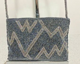 Puede incluir: Un bolso rectangular con cuentas en tonos grises y azules, con un patrón en zigzag en cuentas plateadas e iridiscentes. El bolso tiene una correa de cuerda gris a juego y se exhibe sobre un soporte plateado.