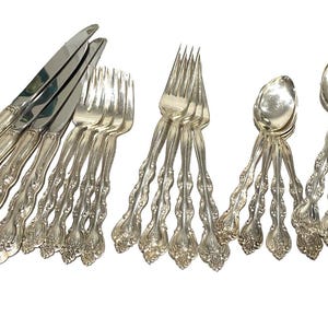 Puede incluir: Un conjunto de cubiertos plateados ornamentados, que incluye cuchillos, tenedores y cucharas. Los mangos presentan un diseño detallado y texturizado. La cubertería está dispuesta sobre una superficie blanca, mostrando la calidad reflectante del metal.