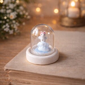 May include: A miniature figurine of a white bunny with pink ears inside a glass dome. The bunny stands on a blue and white cloud, set on a white base. The dome sits on a vintage book, with soft lighting in the background.