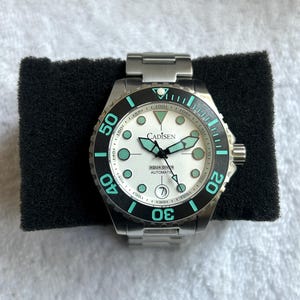 May include: A Cadisen Aqua Diver automatic watch with a silver stainless steel link band and case. The watch features a white dial with luminous teal-colored hour markers and hands, a black rotating bezel with teal numerals, and a date window at six.