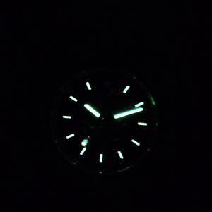 Puede incluir: Primer plano de una esfera de reloj que brilla en la oscuridad. El reloj tiene una esfera negra con manecillas e indicadores verdes luminosos. La hora es aproximadamente las 2:10. El reloj tiene un dise&ntilde;o cl&aacute;sico y redondo.