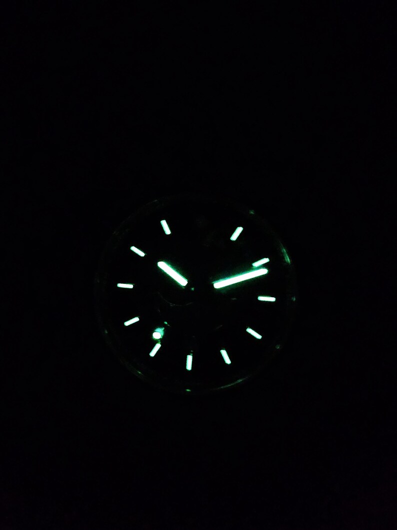 Puede incluir: Primer plano de una esfera de reloj que brilla en la oscuridad. El reloj tiene una esfera negra con manecillas e indicadores verdes luminosos. La hora es aproximadamente las 2:10. El reloj tiene un dise&ntilde;o cl&aacute;sico y redondo.