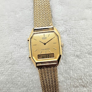 May include: A gold-toned Seiko Quartz watch with a mesh band. The watch face is square with a digital display showing the day and date. The watch has a classic, vintage design.