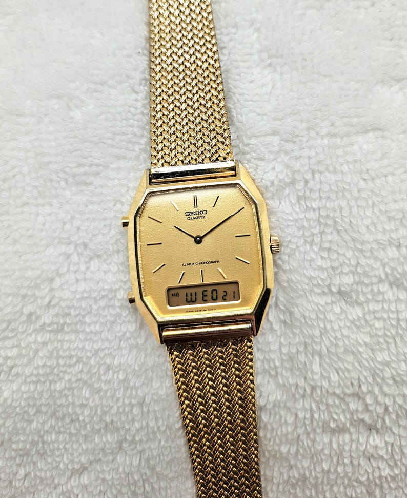 May include: A gold-toned Seiko Quartz watch with a mesh band. The watch face is square with a digital display showing the day and date. The watch has a classic, vintage design.
