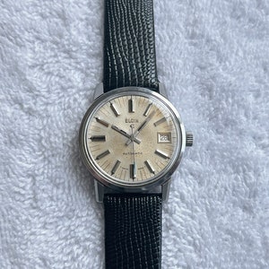 May include: A silver Elgin automatic wristwatch with a white face and black leather band. The watch face has black hour markers and hands, a date window at the 3 o'clock position, and the words "Elgin Automatic" printed below the center.