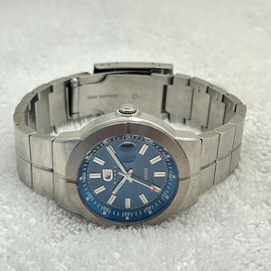 May include: A stainless steel Guess watch with a blue face. The watch has a silver strap and a blue bezel. The watch face features the Guess logo and white hour markers. The watch is displayed on a white surface.