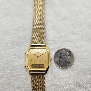 May include: A gold-toned Seiko Quartz alarm chronograph watch with a mesh band. The watch face is rectangular with a digital display showing the date. A US quarter is next to the watch for size comparison.